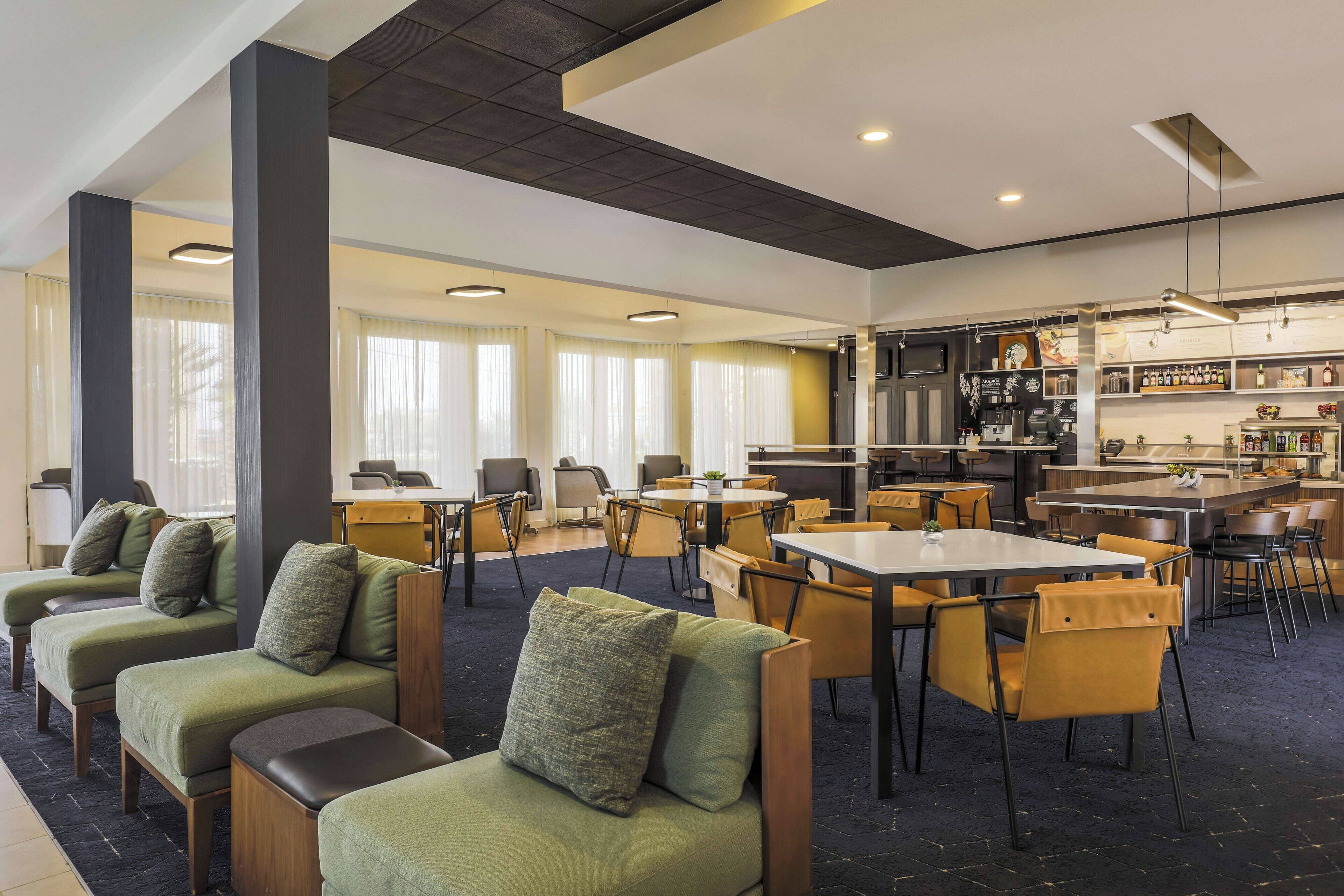 Un moderno vestíbulo de hotel con sofás verdes, sillas amarillas, una zona de comedor central y una cocina al fondo.