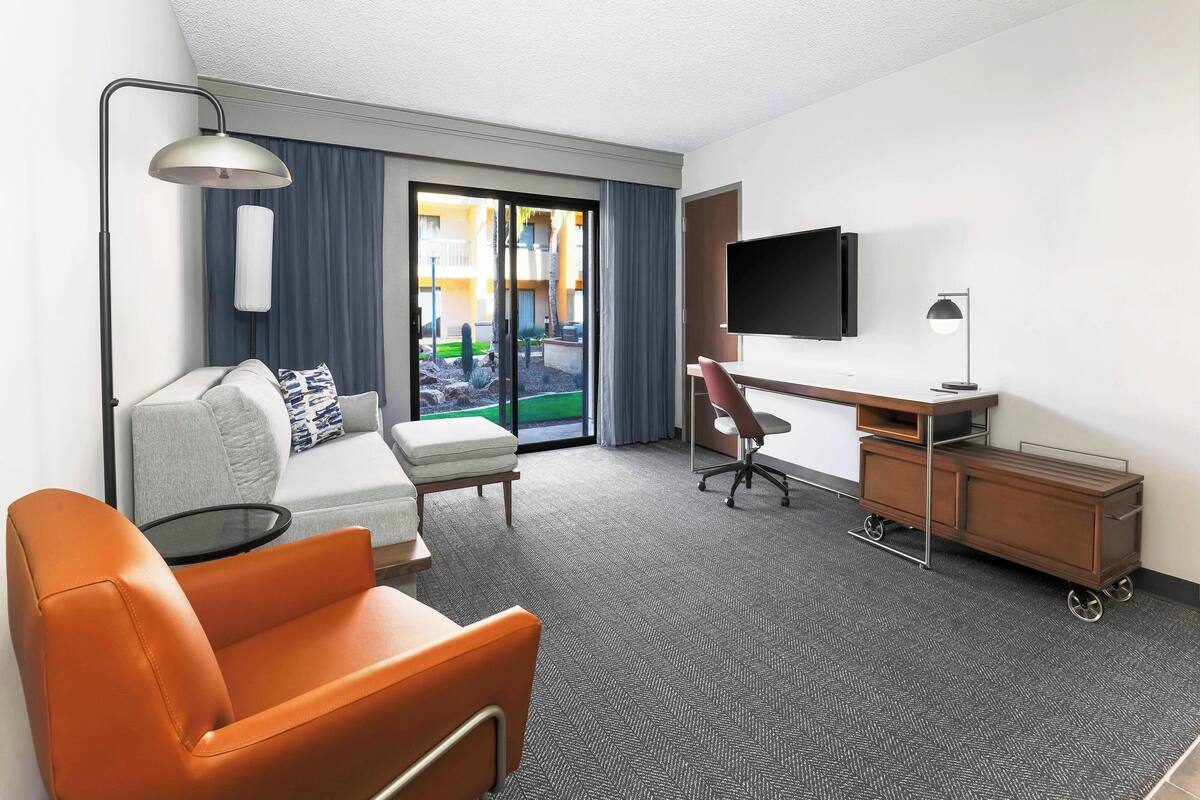 A hotel room with a sofa, an orange chair, a desk, a TV, and a view of a courtyard.