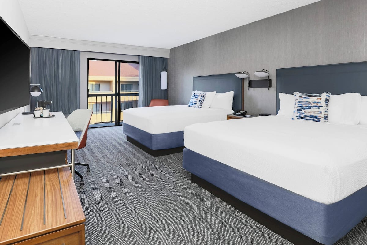 A hotel room with two beds, a desk, a chair, and a view of the outside.
