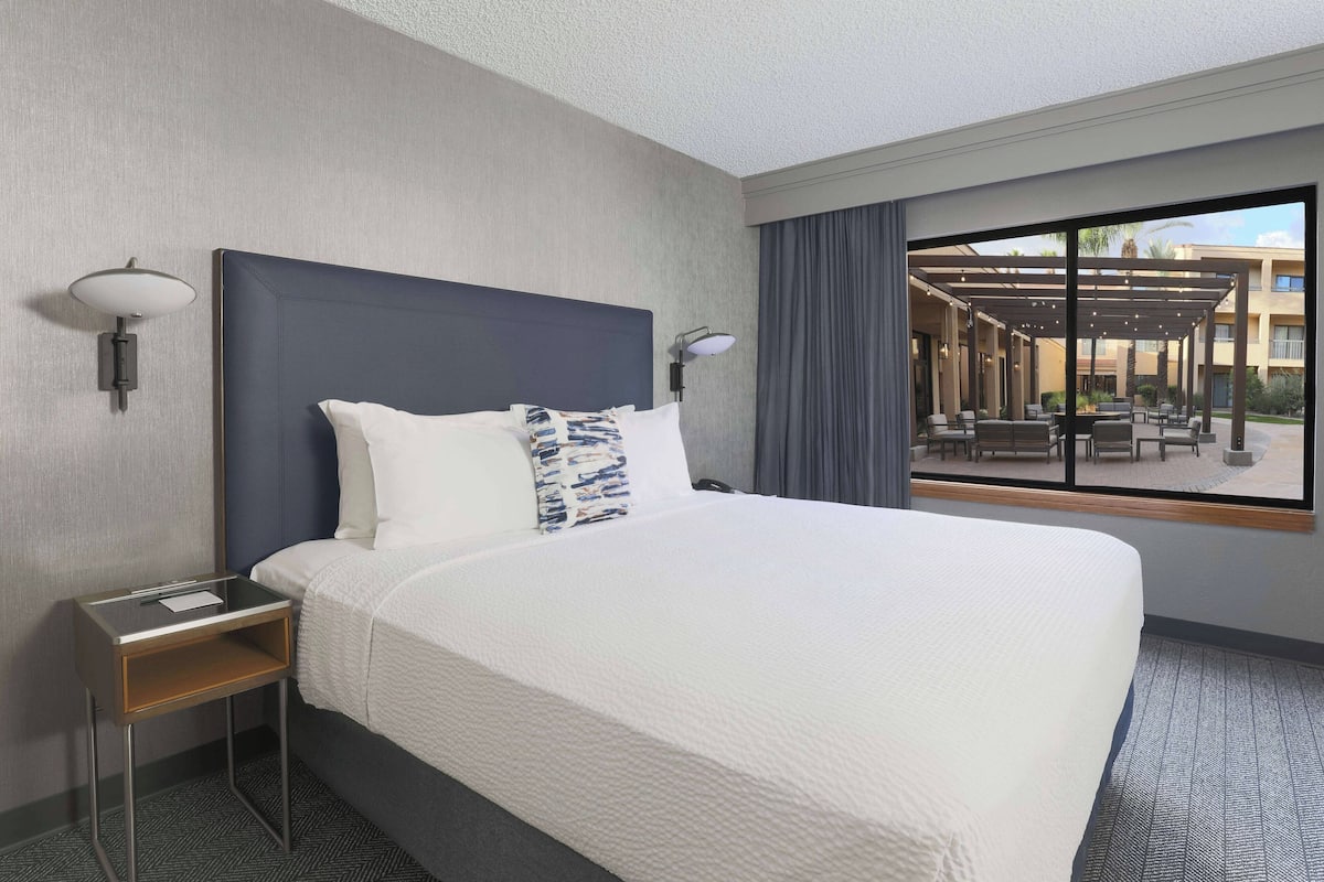 A hotel room with a large bed, a nightstand, two wall-mounted lamps, and a view of the outdoor patio through a large window.