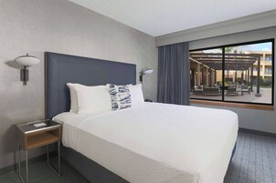 Quarto de hotel com uma cama grande, uma mesa de cabeceira, duas luminárias fixadas na parede e vista para o pátio externo através de uma janela ampla.