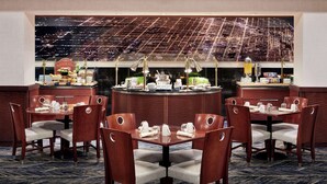 En restaurang med ett stort, runt bord i mitten, omgivet av stolar, en buffédisk med mat och dryck samt ett stort fönster med utsikt över staden.