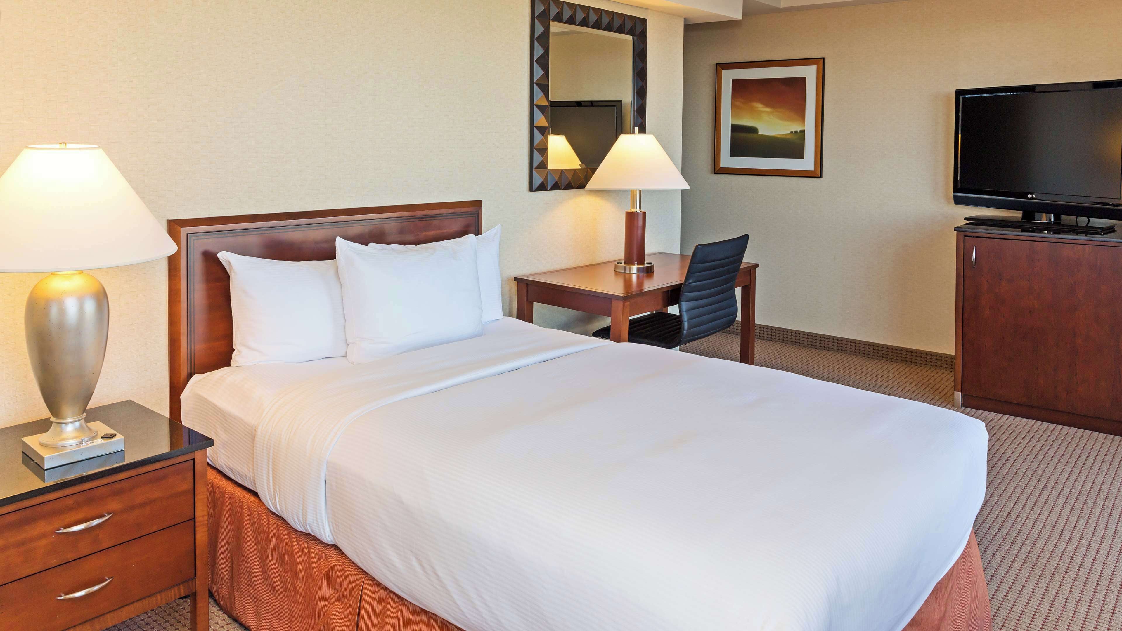 A hotel room with a bed, two bedside lamps, a desk with a chair, a television, and a framed picture on the wall.