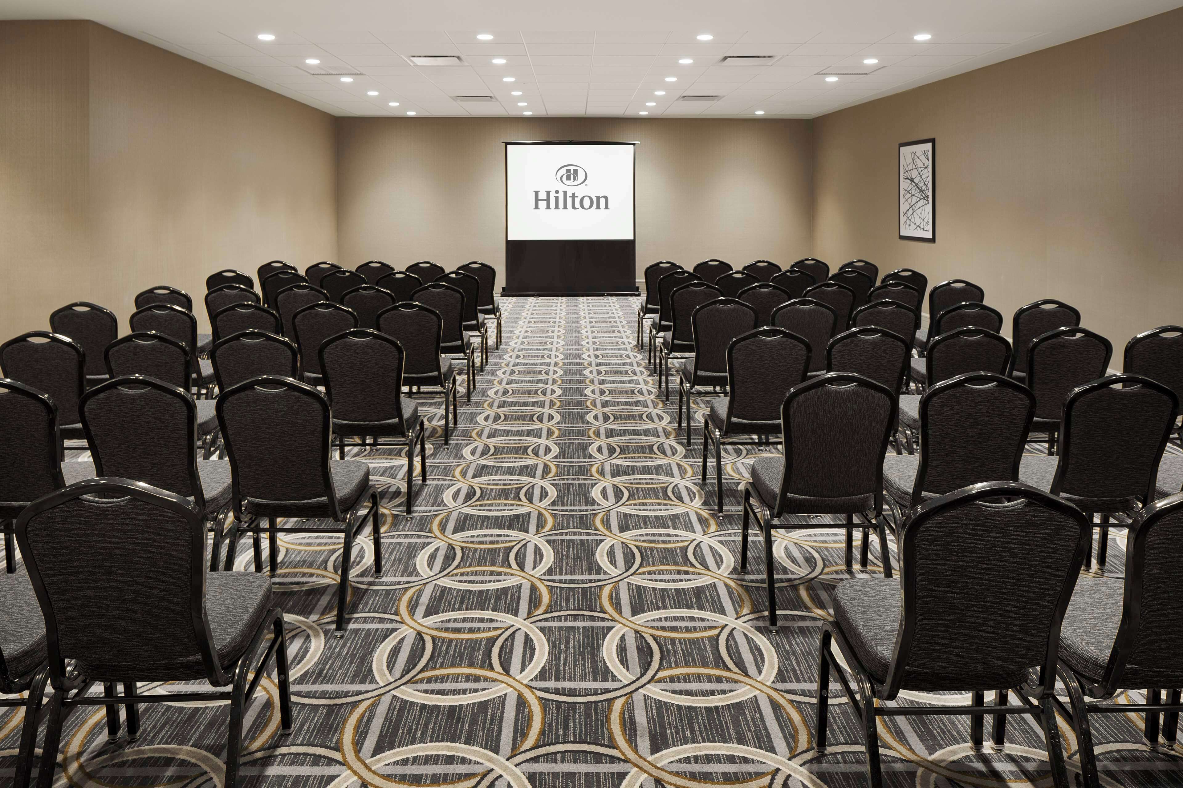 Ein Konferenzraum mit Reihen von Stühlen, einem Teppichboden und einer Projektionsleinwand mit dem Hilton-Logo.