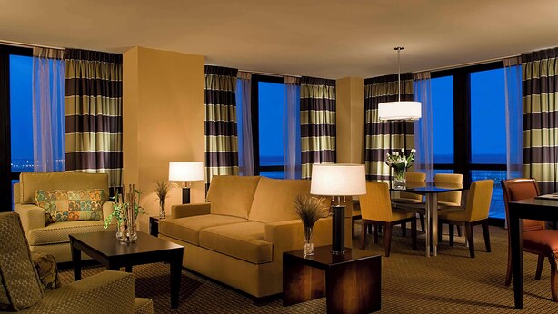 Habitación de hotel con sofá, sillones, una mesa de comedor y vistas de la ciudad de noche.