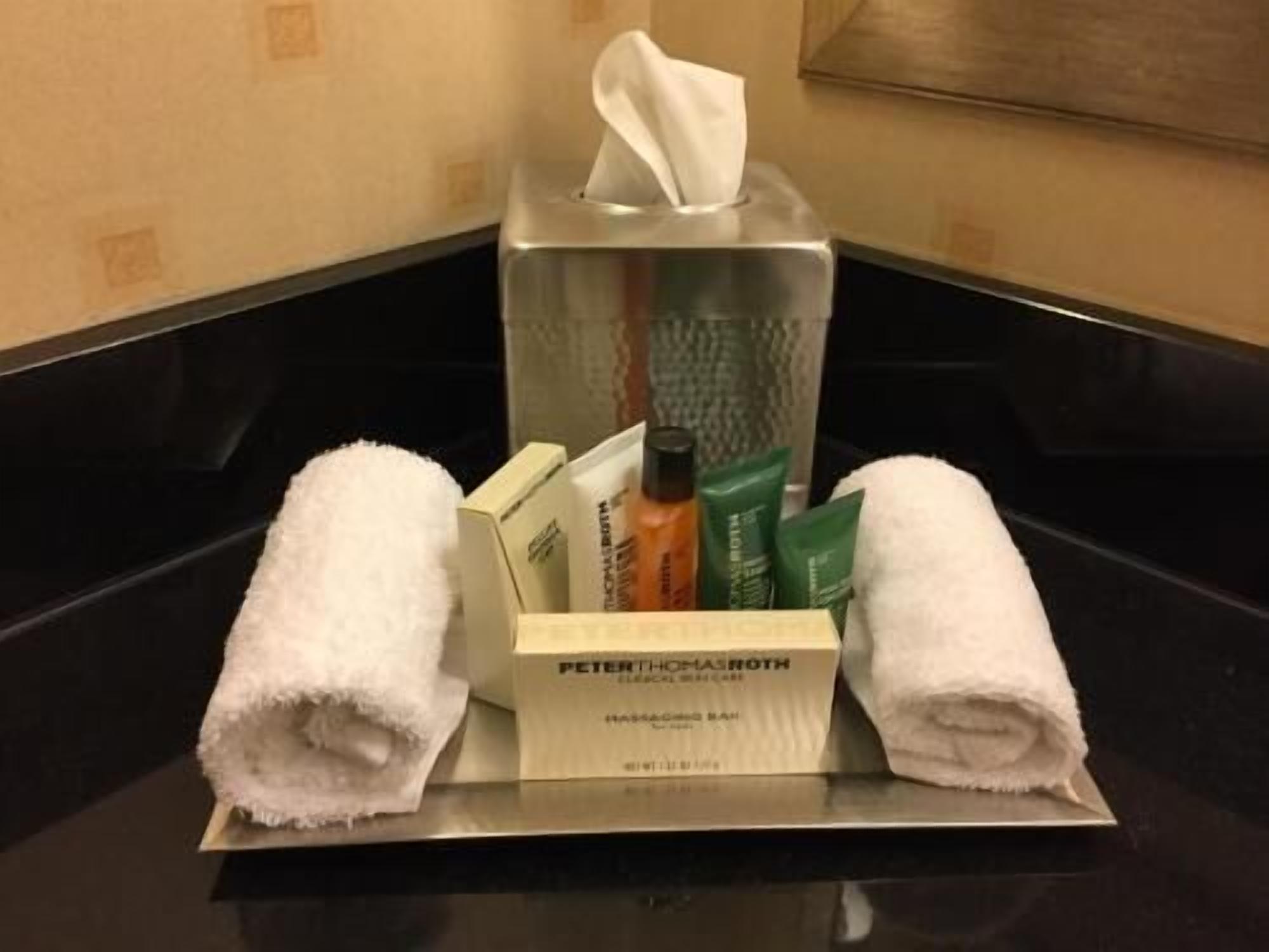 Une chambre d’hôtel avec un meuble-lavabo dans la salle de bain, une boîte de mouchoirs, des articles de toilette et deux serviettes enroulées.