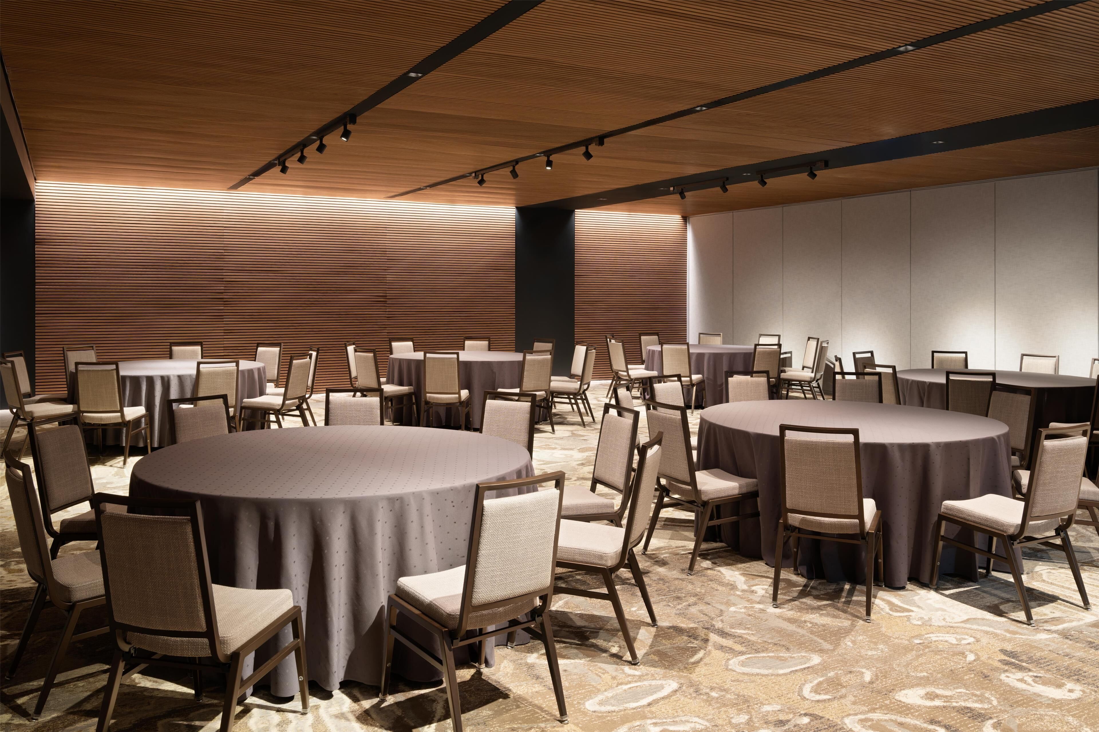 A conference room with multiple round tables and chairs, wooden panel walls, and a carpeted floor.