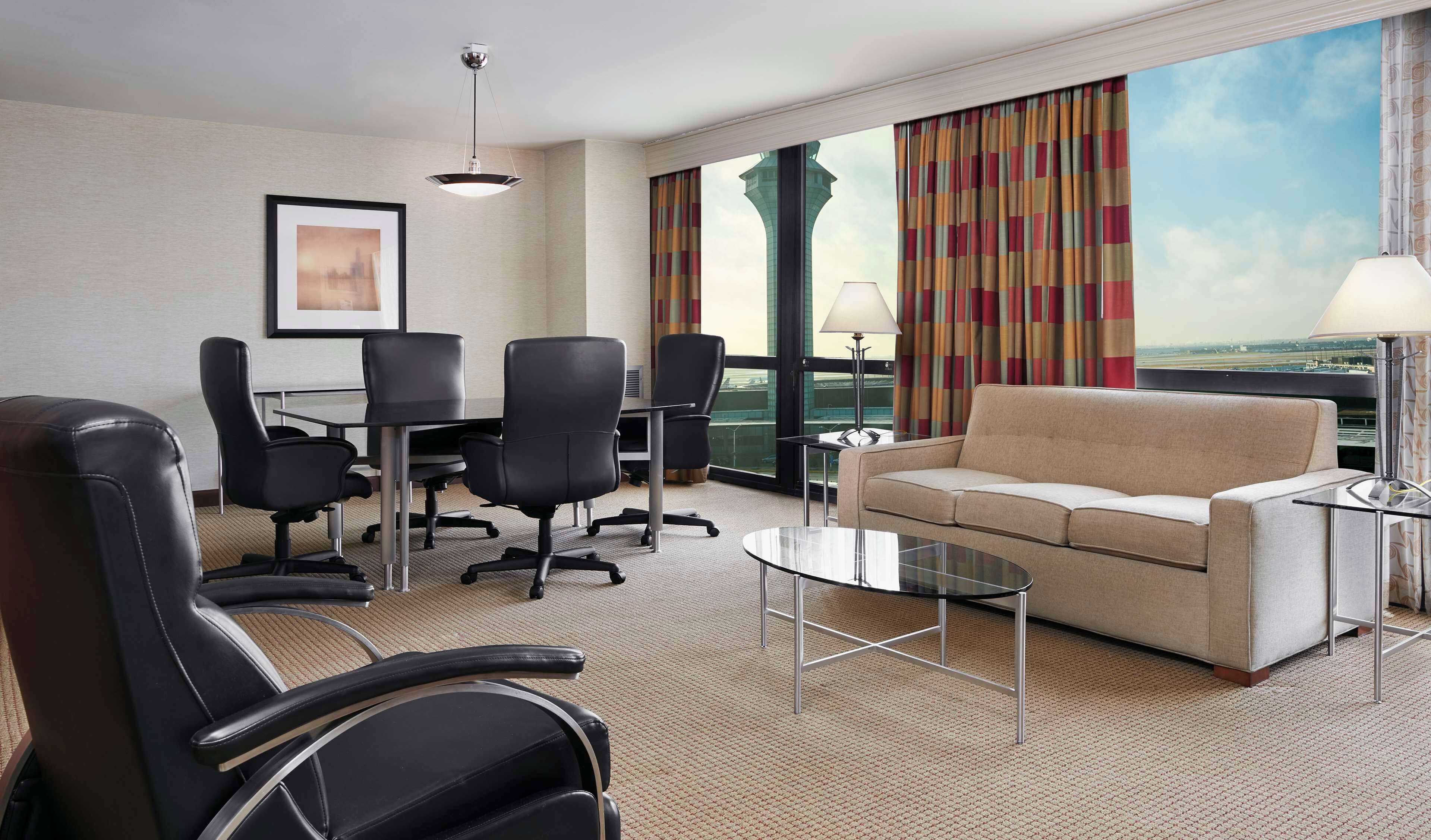 A hotel room with a large window, a sofa, a coffee table, and a conference table with chairs.