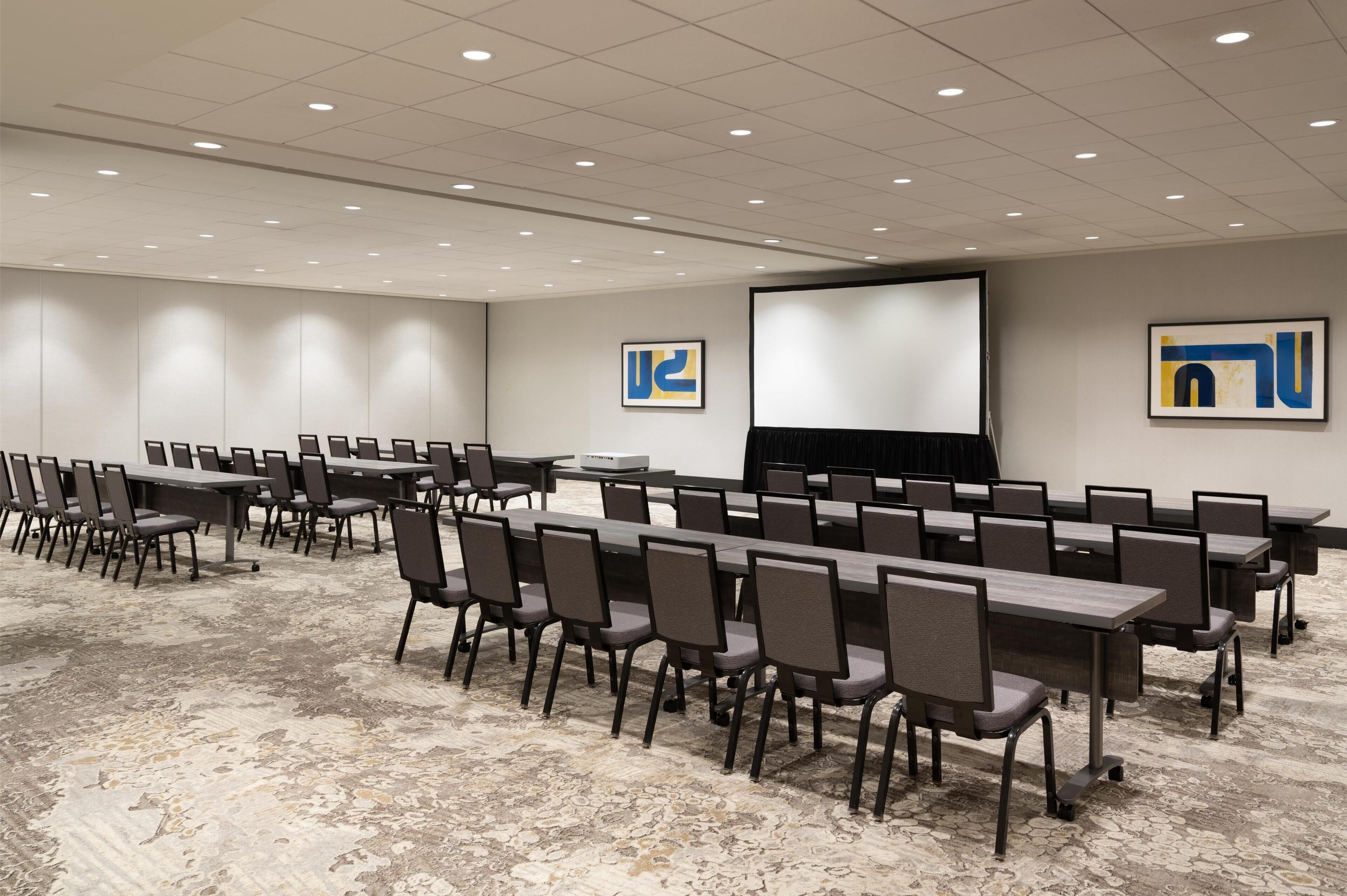 A conference room with rows of tables and chairs, a projector screen, and two abstract paintings on the walls.
