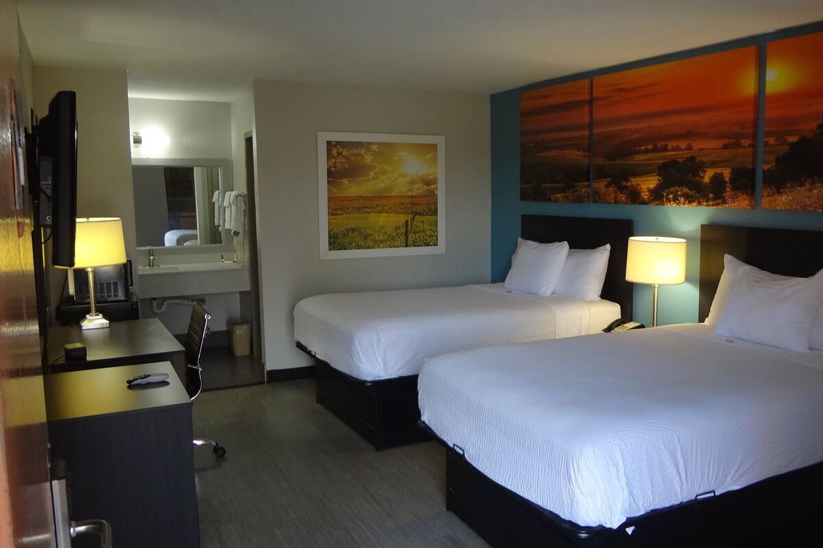 A hotel room with two beds, a desk, a mirror, and artwork on the wall.