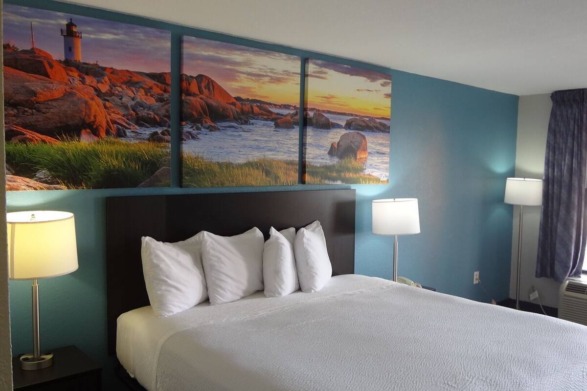 A hotel room with a large bed, two bedside lamps, a headboard, and a wall mural of a coastal scene.