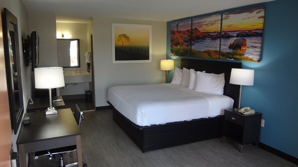 A hotel room with a large bed, two bedside tables with lamps, a desk with a chair, a bathroom with a mirror and sink, and a painting on the wall.