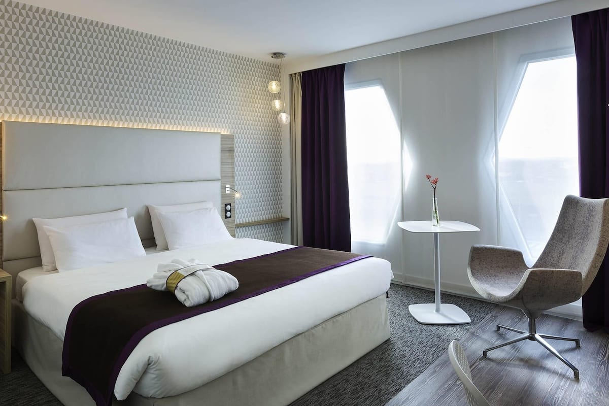 A hotel room with a large bed, a small round table, a chair, and a view of the outdoors.