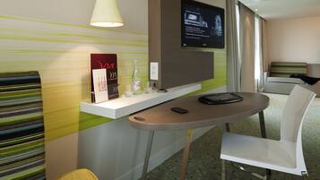 A hotel room with a modern design, featuring a small table with a chair, a television mounted on the wall, and a window with curtains.