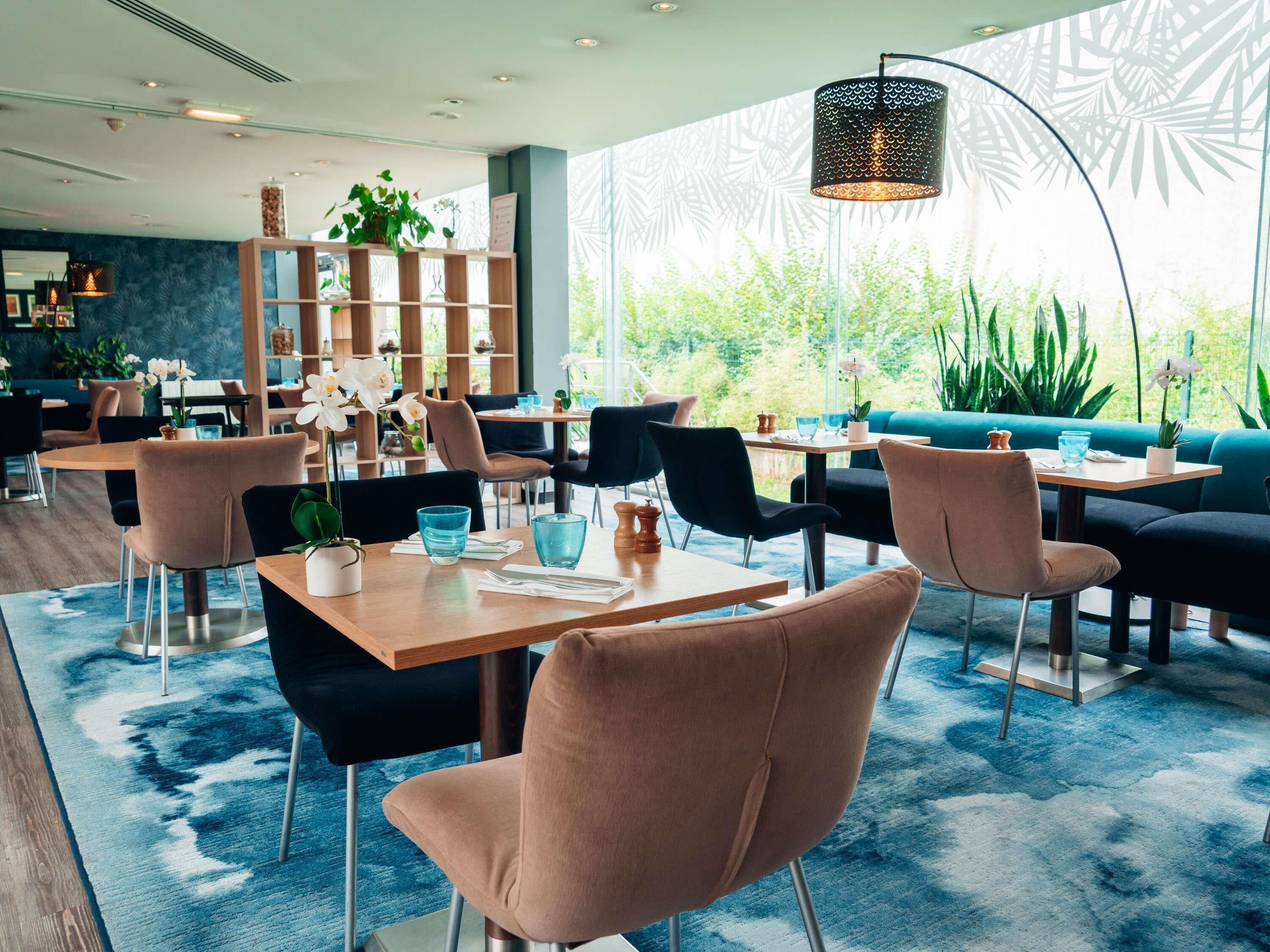 Un restaurant moderne avec des tables rondes, des chaises rembourrées et une grande fenêtre donnant sur un paysage verdoyant.