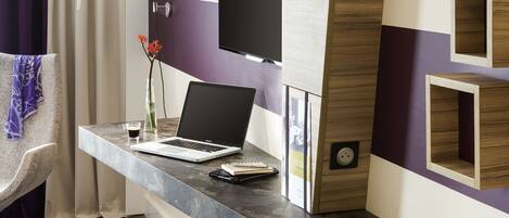 A modern hotel room with a desk, laptop, chair, and a large TV on the wall.