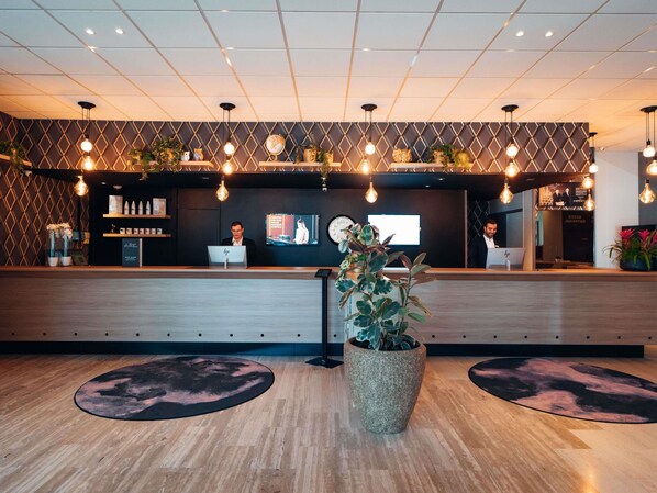 A reception area with a wooden reception desk, two receptionists, a clock, and a large potted plant.