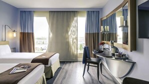 A hotel room with two beds, a desk, a chair, a large mirror, and a view of the city.
