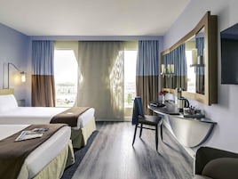 A hotel room with two beds, a desk, a chair, a large mirror, and a view of the city.