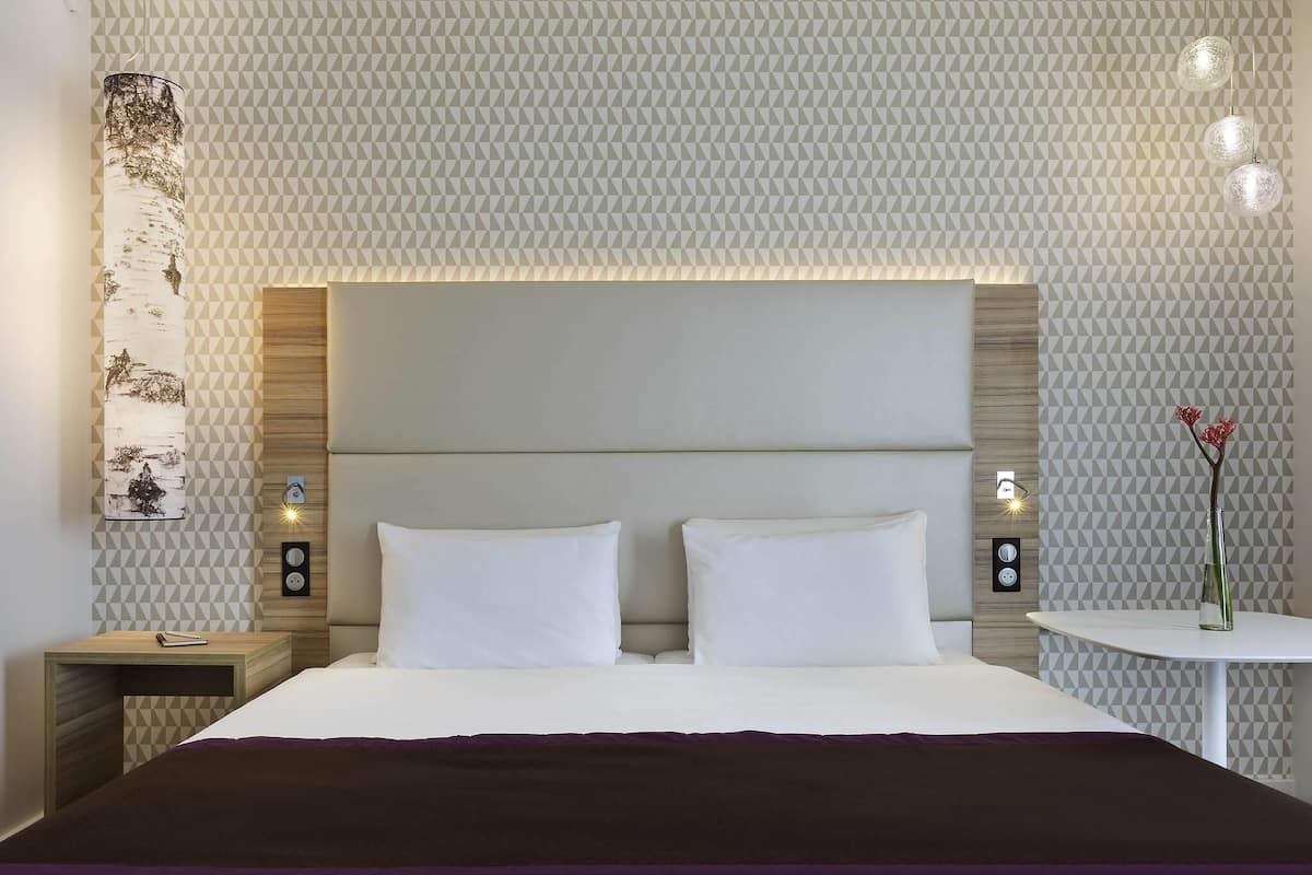 A hotel room with a large bed, a bedside table, a nightstand, a wall-mounted light, and a small table with a vase.