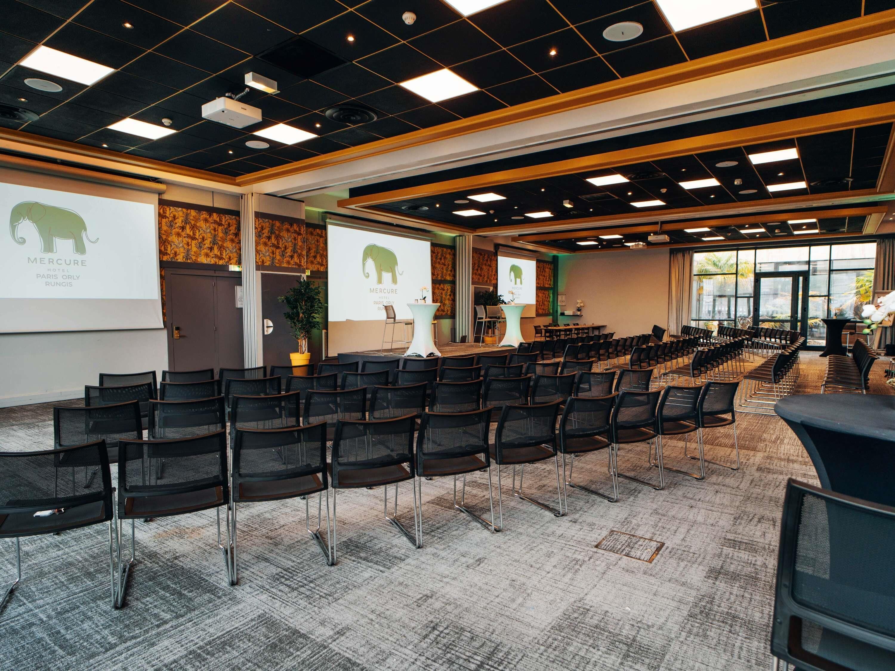 Een congrescentrum met rijen stoelen, een podium en presentatieschermen waarop de naam van het hotel staat.