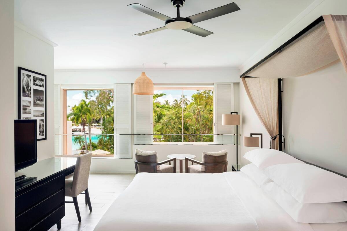 Habitación de hotel moderna con cama, escritorio y sillas, ventanas amplias con vistas a palmeras y una piscina, además de un ventilador de techo.