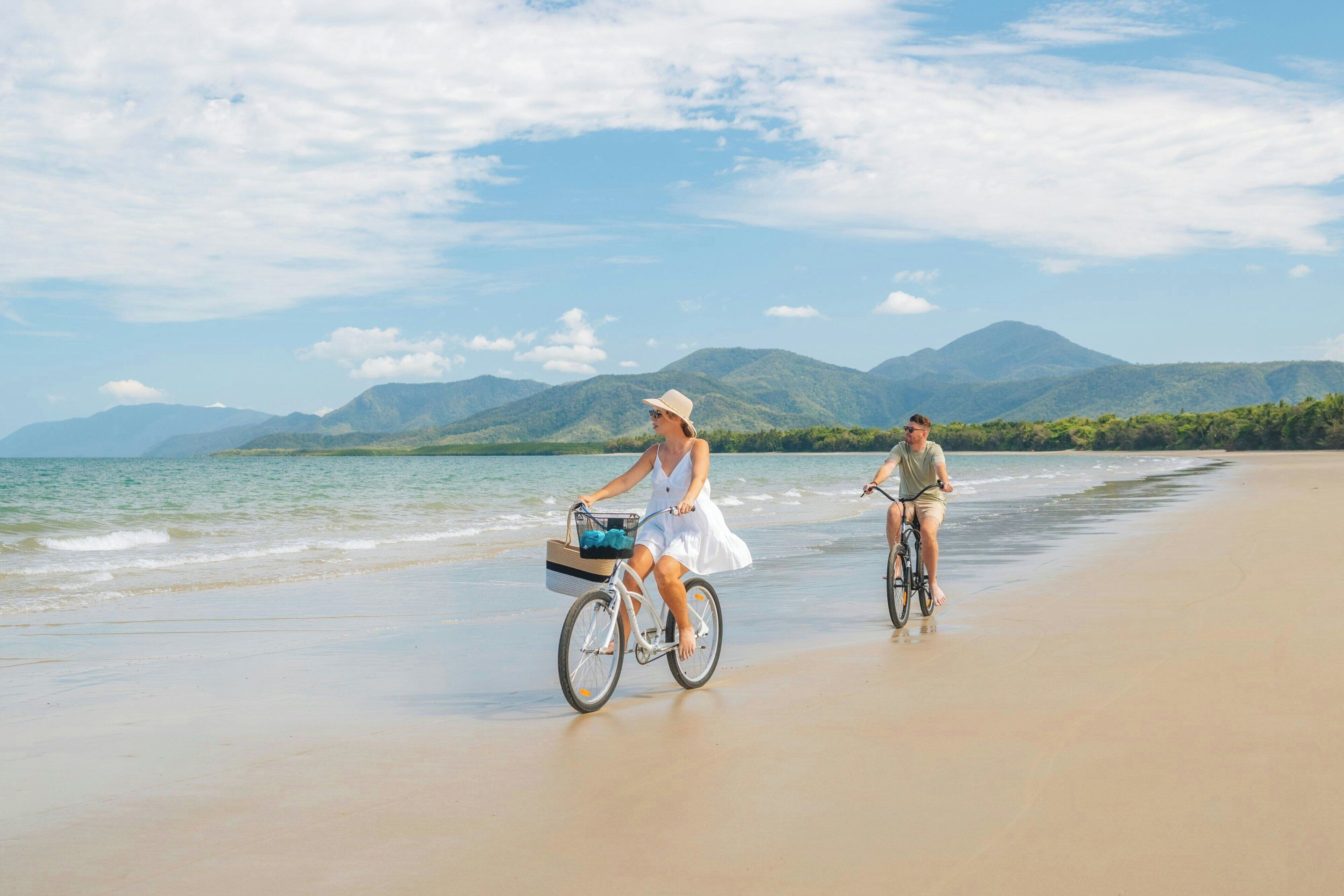 Twee personen fietsen op het strand, met bergen op de achtergrond.