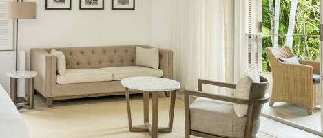 A modern hotel room with a beige sofa, a round table, a wicker chair, and a view of greenery through large windows.