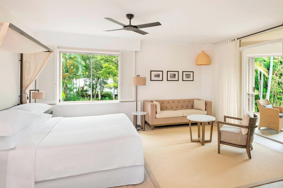 Habitación de hotel con cama, sofá, sillón, una mesita y un ventilador de techo. Hay ventanas con cortinas, una lámpara y cuadros en la pared.