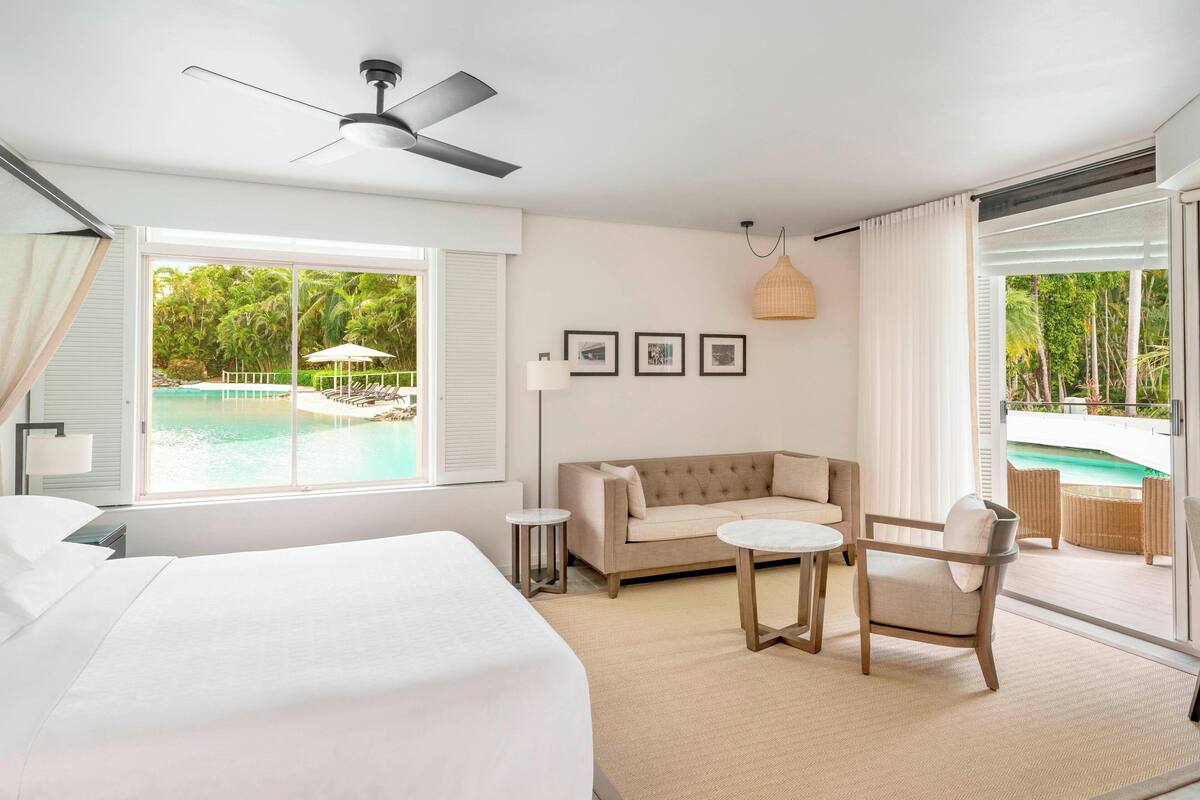 Habitación de hotel con cama, sofá, una mesita y una silla. Se ve una ventana con vista a una piscina y palmeras.