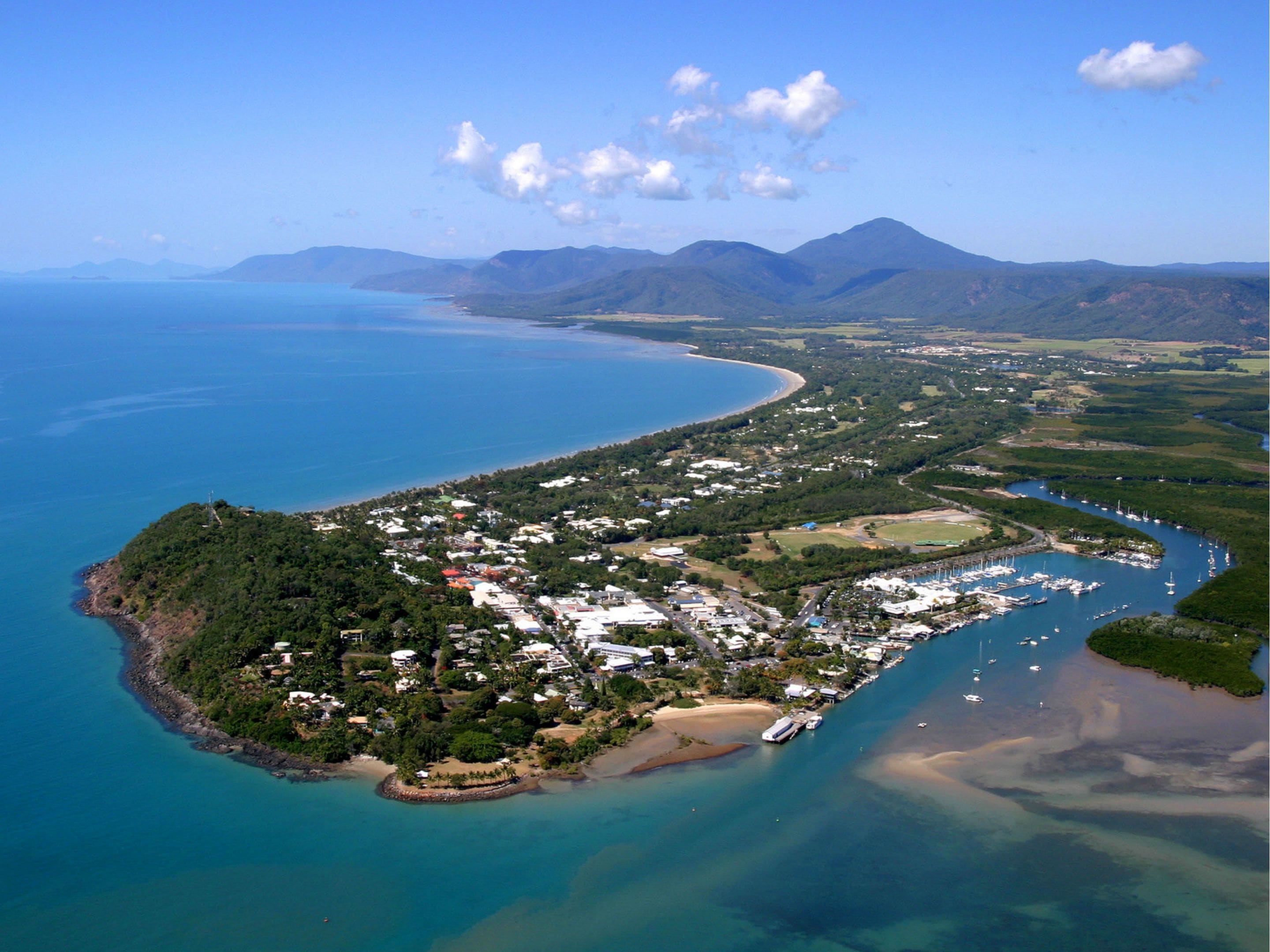 An aerial view of a coastal town with a marina, residential areas, and surrounding mountains.