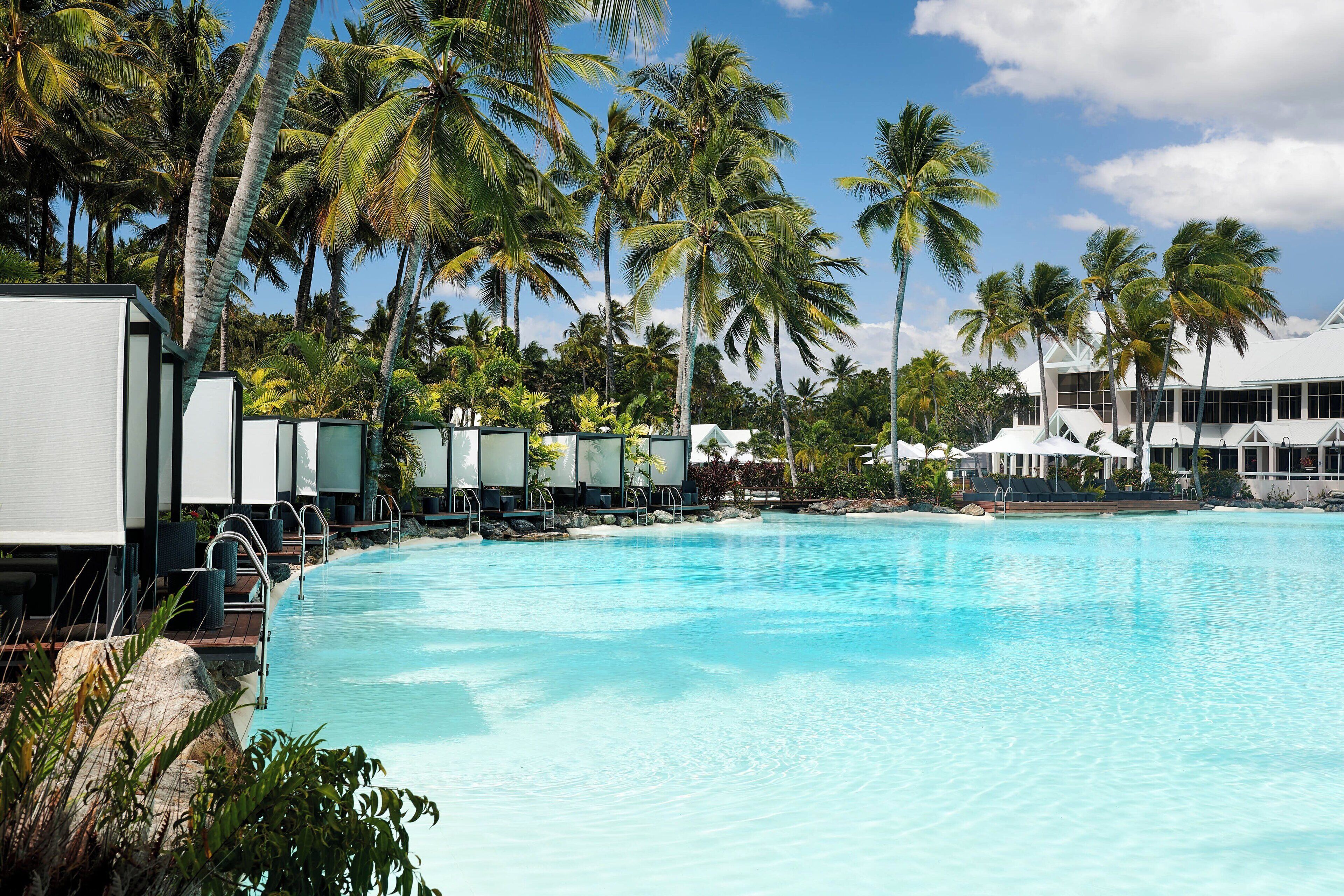 A large swimming pool with lounge chairs and umbrellas, surrounded by palm trees and a resort building.