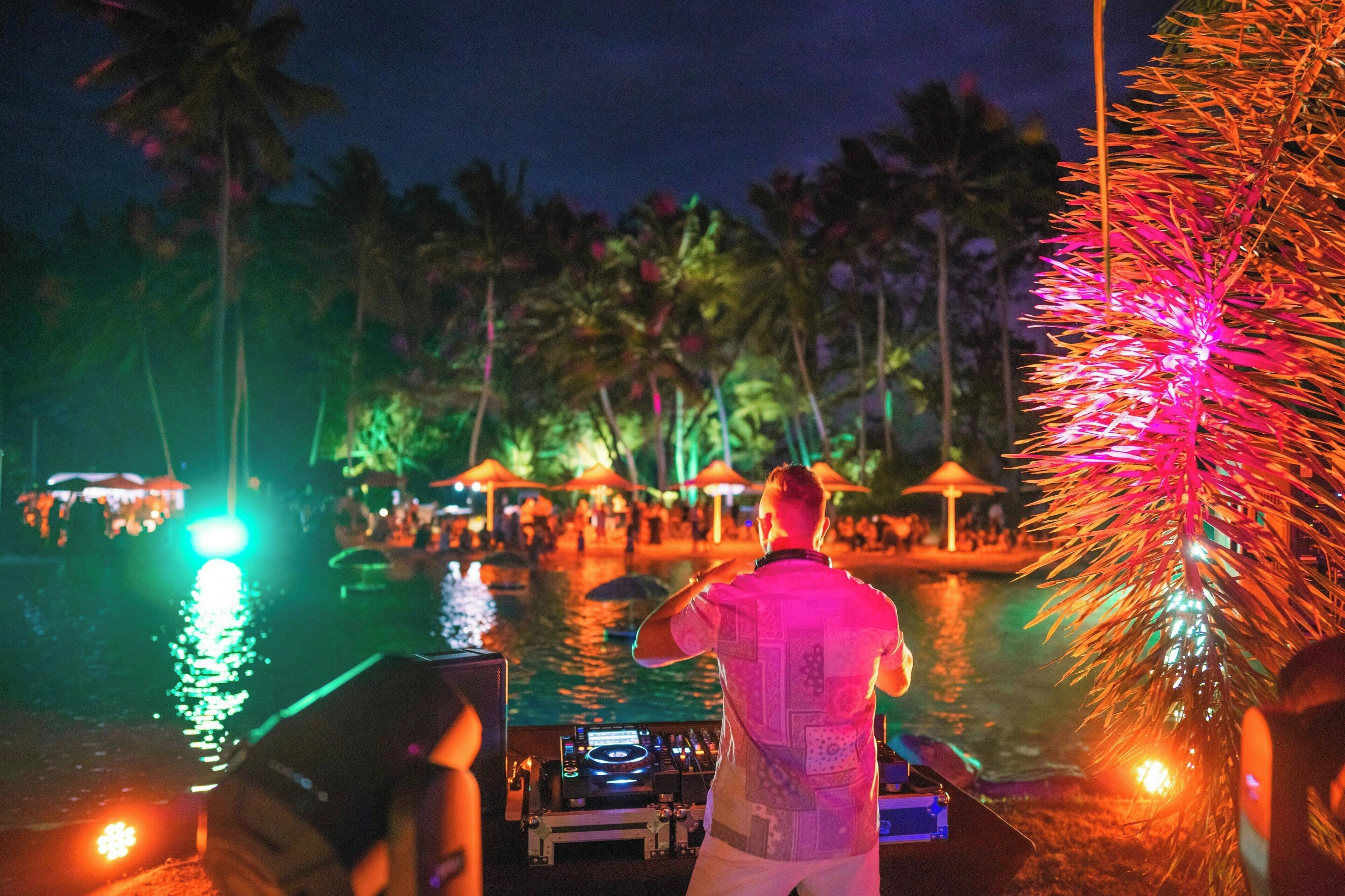 A person standing by a pool with palm trees and umbrellas in the background.