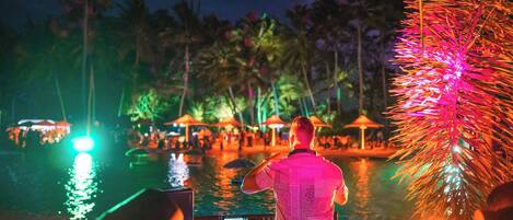 A person standing by a pool with palm trees and umbrellas in the background.