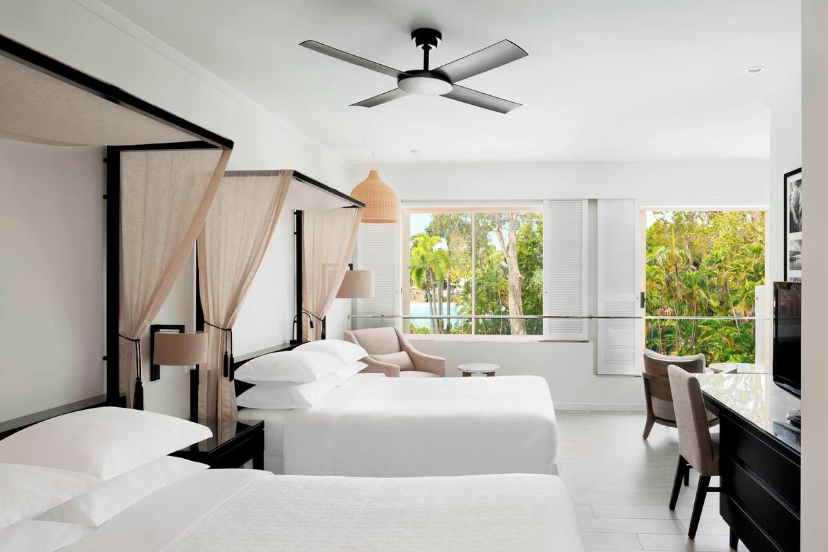Habitación de hotel moderna con dos camas, ventilador de techo, ventana grande con vistas a zonas verdes y un escritorio con silla.