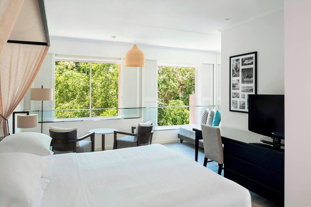 Habitación de hotel con una cama grande, un escritorio, una silla, un televisor y un balcón con vistas a zonas verdes.