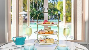 A three-tiered dessert stand with various pastries and sandwiches, two glasses of champagne, a blue teapot, and cups on a table with a view of a pool and palm trees. - Sheraton Grand Mirage Resort, Port Douglas (Port Douglas)