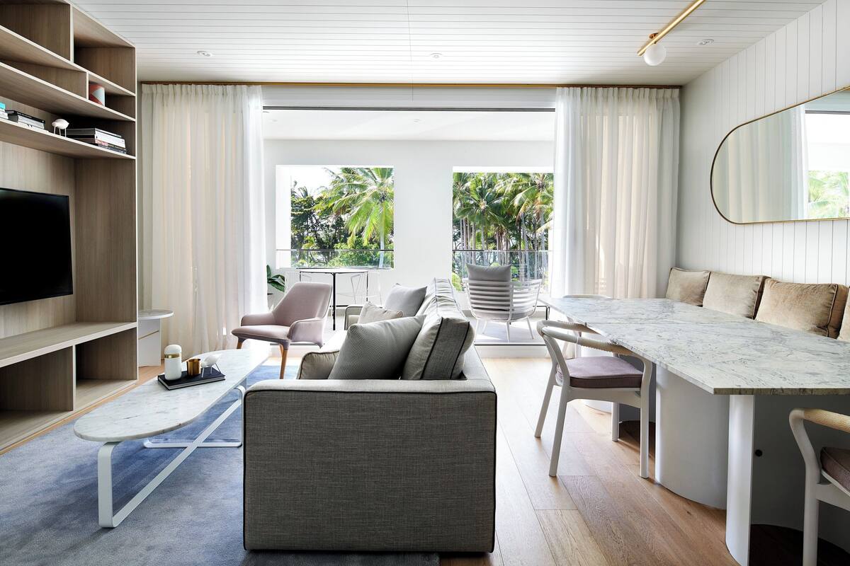 Una sala de estar moderna con un sofá, una mesa de centro y una mesa de comedor. Hay un televisor a la izquierda y un balcón con vistas a palmeras.