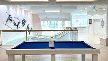 Ein Billardtisch in einem geräumigen, gut beleuchteten Raum mit modernem Design, großen Fenstern und Blick auf einen Poolbereich.