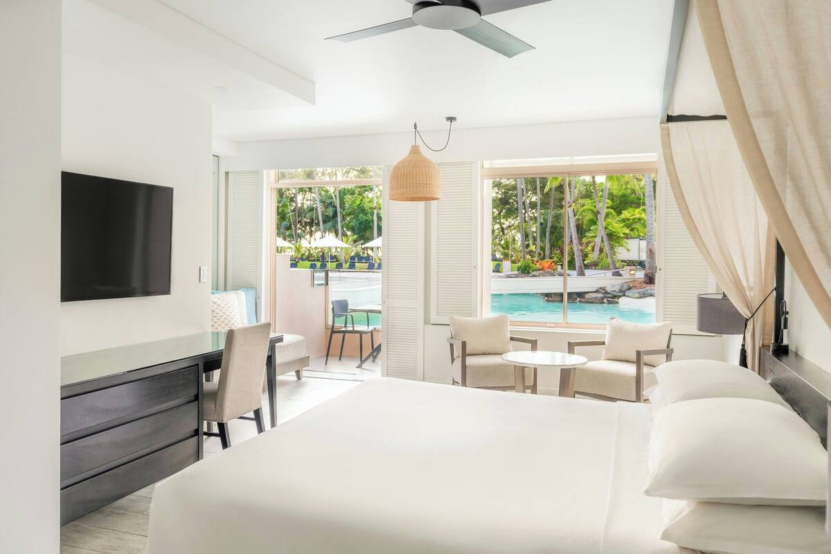 Una habitación de hotel moderna con una cama grande, un televisor, un comedor con mesa y sillas, y vistas a una piscina y paisaje tropical.