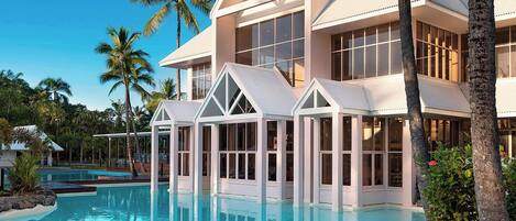Un resort con una piscina grande, palmeras y un edificio moderno con ventanales amplios.
