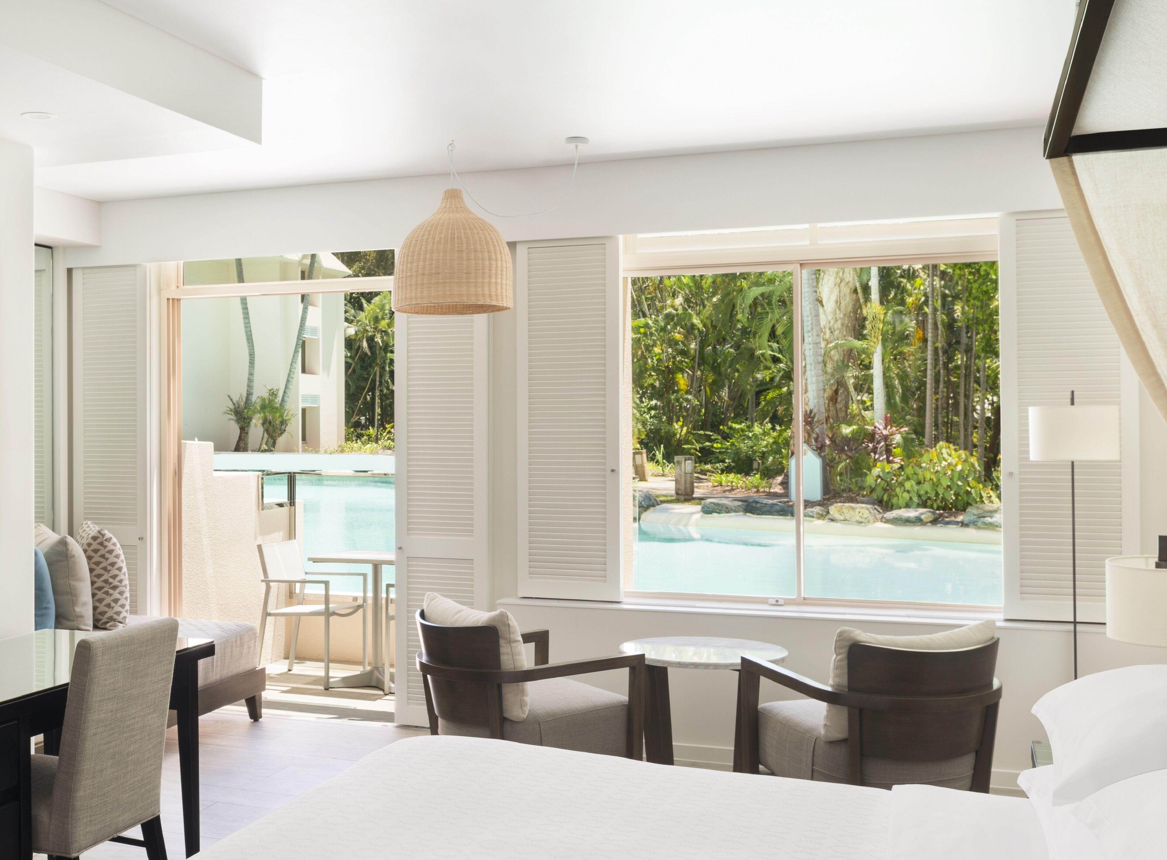 A hotel room with a bed, a dining table, and a view of a swimming pool and tropical garden.