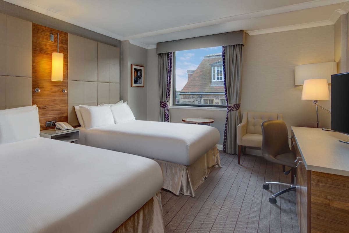 Habitación de hotel con dos camas, un escritorio, una silla, una mesita y una ventana con cortinas.