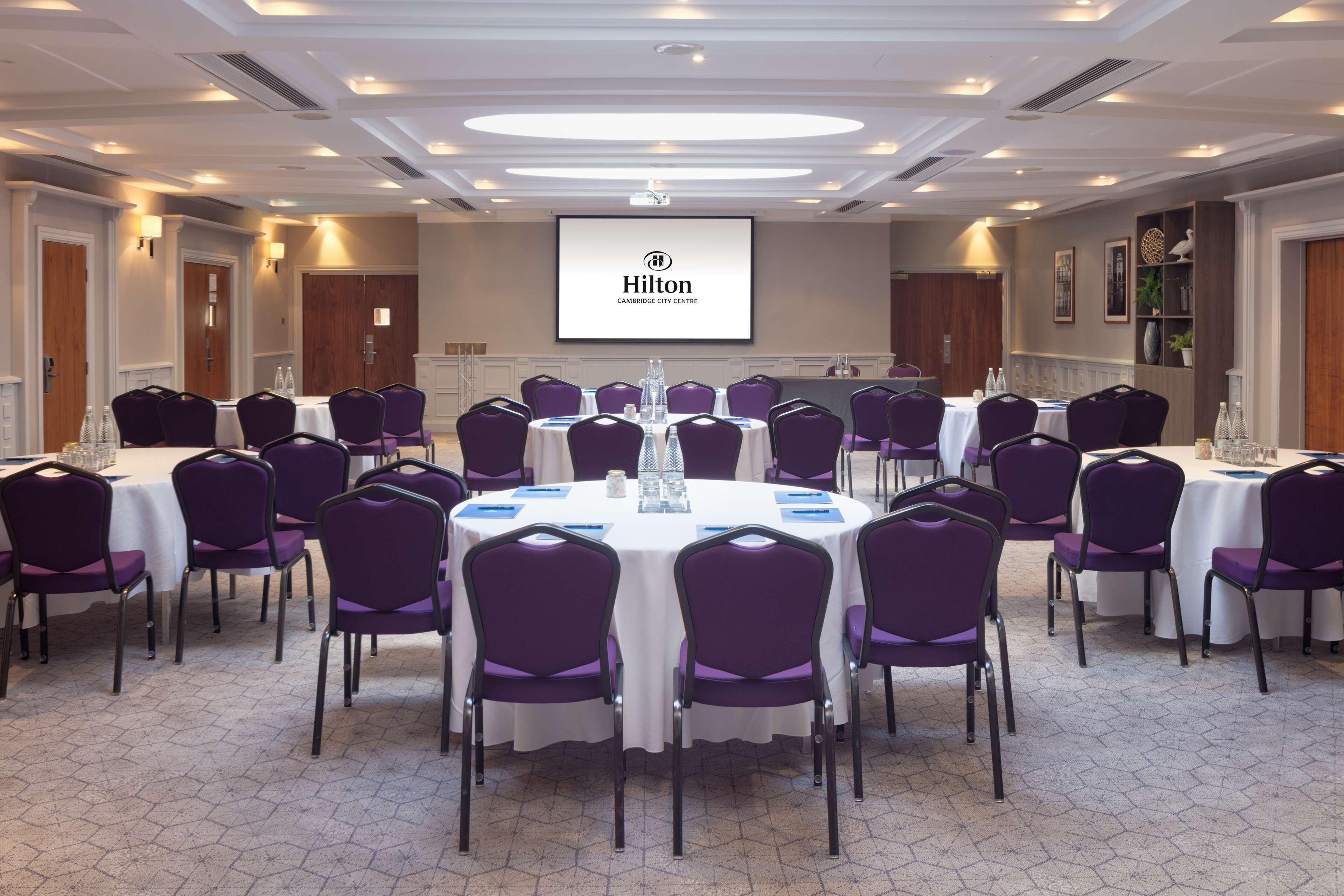 A conference room with round tables, purple chairs, and a projector screen displaying the Hilton Cambridge City Centre logo.
