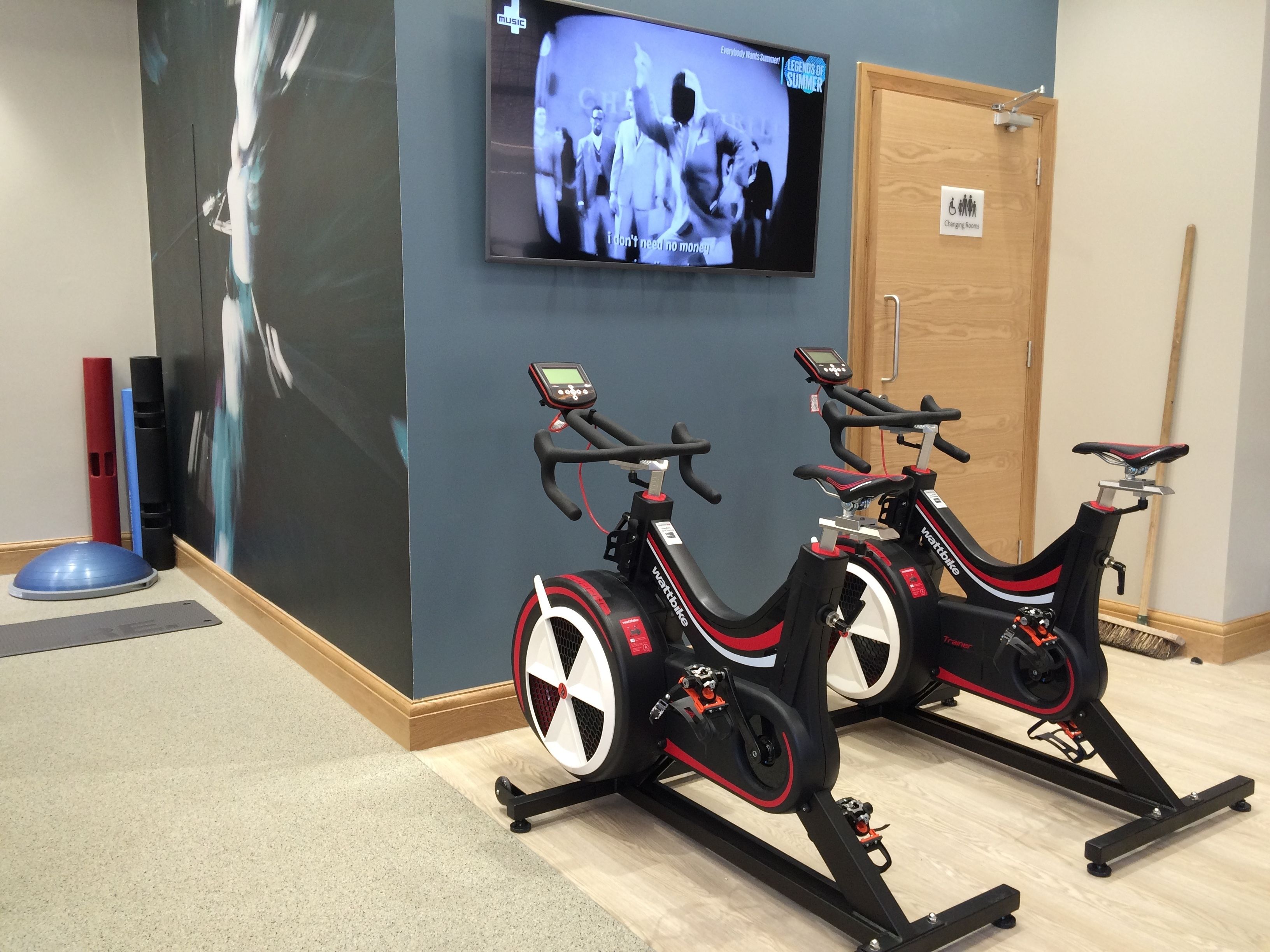 Dos bicicletas estáticas en una habitación con un televisor en la pared mostrando una película.