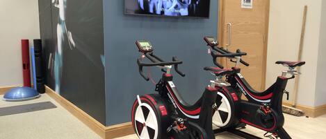 Dos bicicletas estáticas en una habitación con un televisor en la pared mostrando una película.