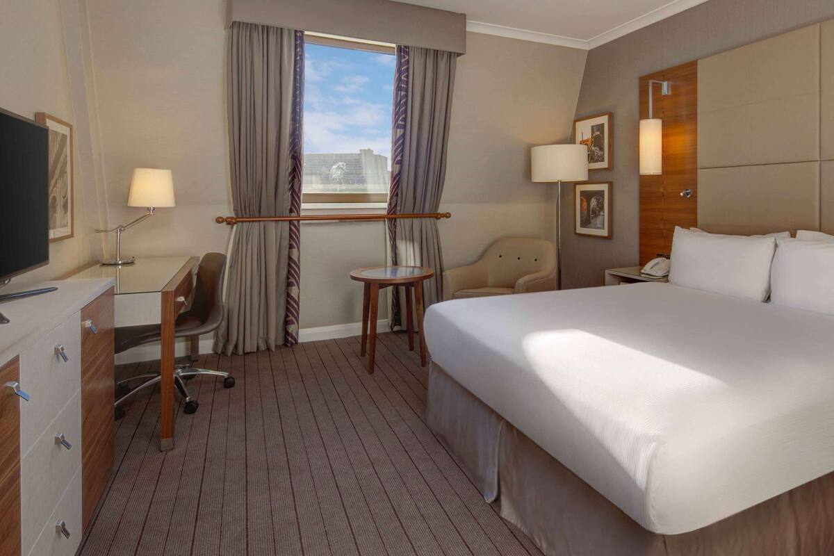 Habitación de hotel con una cama grande, un escritorio con silla, una mesita, televisión y ventana con cortinas.