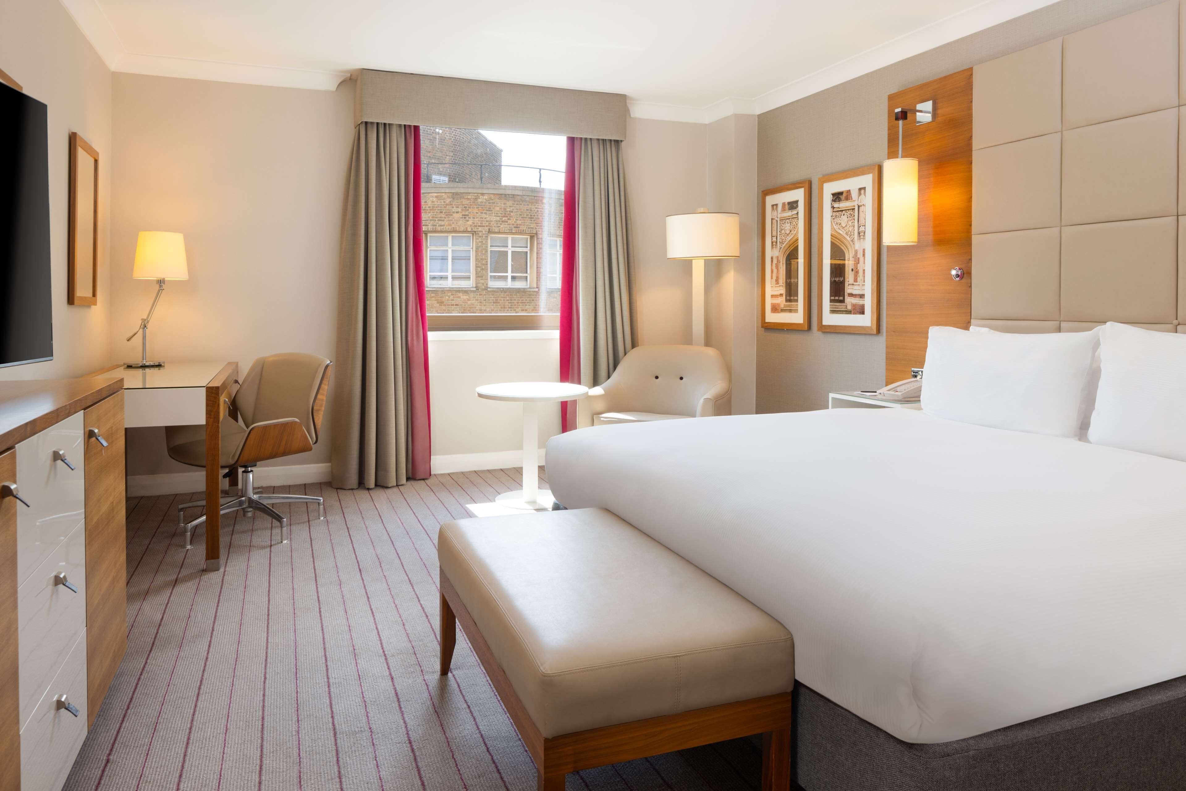A hotel room with a large bed, a desk with a chair, a small table, a TV, and a window with curtains.