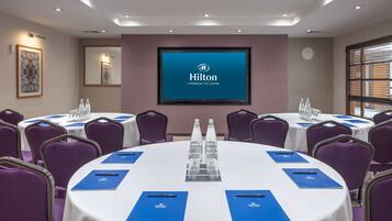 A conference room with round tables, purple chairs, and a large screen displaying the Hilton logo.