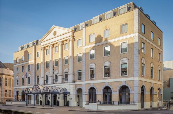 A multi-story hotel with a classical facade, featuring a central entrance with a covered walkway, large windows, and a flagpole. - Hilton Cambridge City Centre (Cambridge)